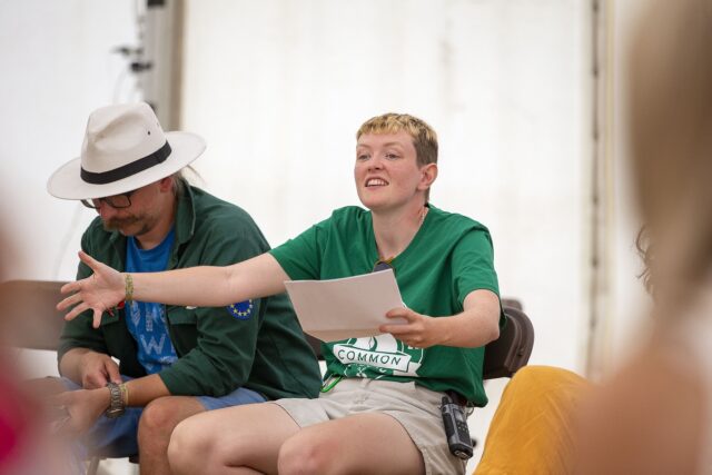 A young person with blonde hair and a green t-shirt reads out a story in a tent as part of their work with the Woodcraft Folk charity