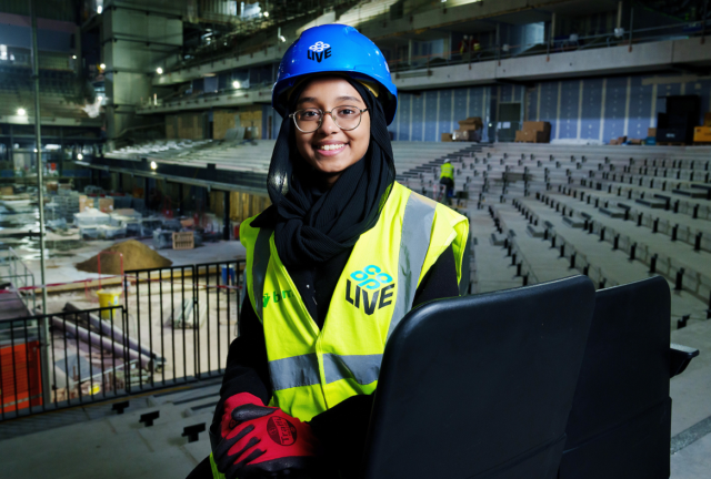 Binish Syed Qureshi sat on the first seats installed at Co-op Live. She's wearing a high-vis jacket and a hard hat that say Co-op Live on them. 