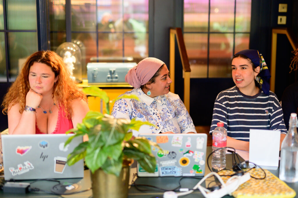 “Three people sit at a table working on laptops covered in colorful stickers in a bright, modern workspace.”