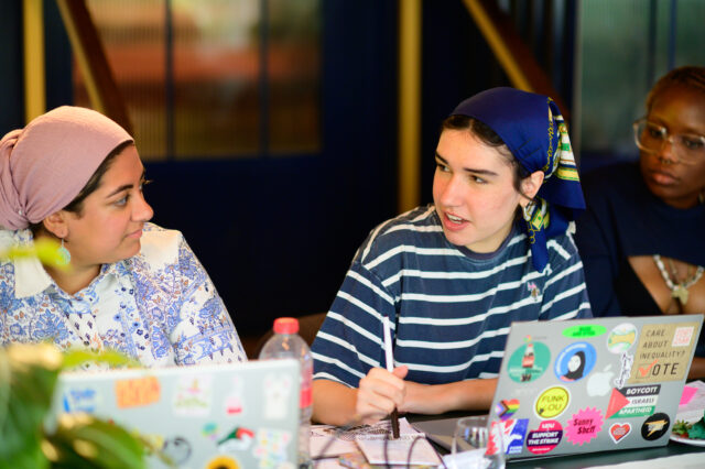 Three young people from our Future Communities Collective, sitting at a table engaged in collaborative work. Two open laptops covered in colorful stickers are visible on the table, along with notebooks, pens, and a water bottle. The individuals are wearing patterned headscarves and casual clothing.