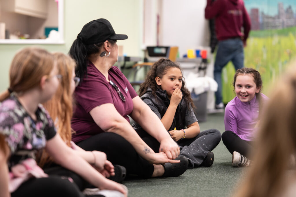 A photo of one of a session at Re:establish , one of our partners funded by our Future Community Fund. There's a woman with a black cap sat crossed legged on the floor, with four girls sat next to her. 
