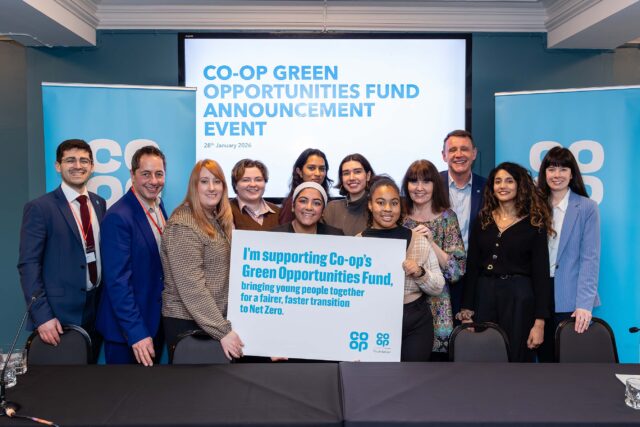 A group of people standing together at an indoor event, holding a large printed sign that reads: ‘I’m supporting Co-op’s Green Opportunities Fund, bringing young people together for a fairer, faster transition to Net Zero.’ Behind them is a presentation screen displaying the title ‘Co-op Green Opportunities Fund Announcement Event – 28th January 2026.’ Two Co-op–branded banners stand on either side of the group, and a long table with microphones is positioned in the foreground.