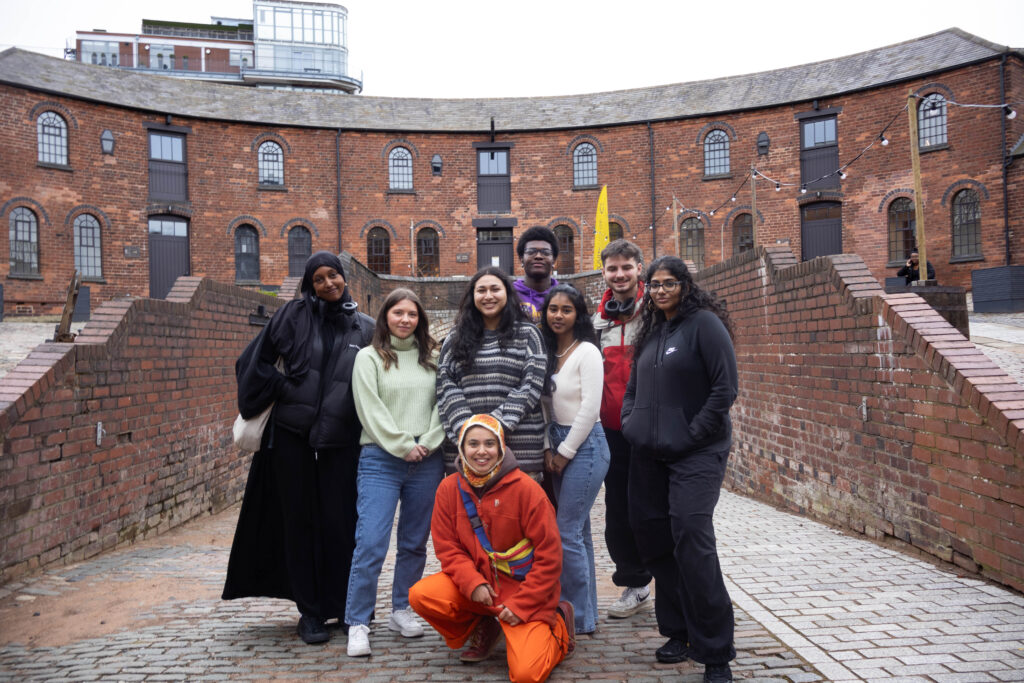 A group of 8 young people stood together smiling at the camera in front of a red brick building.