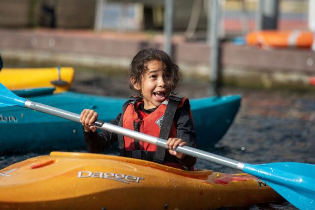 A young girl is in an orange kayak in a canal in west London. She's smiling and holding a blue oar