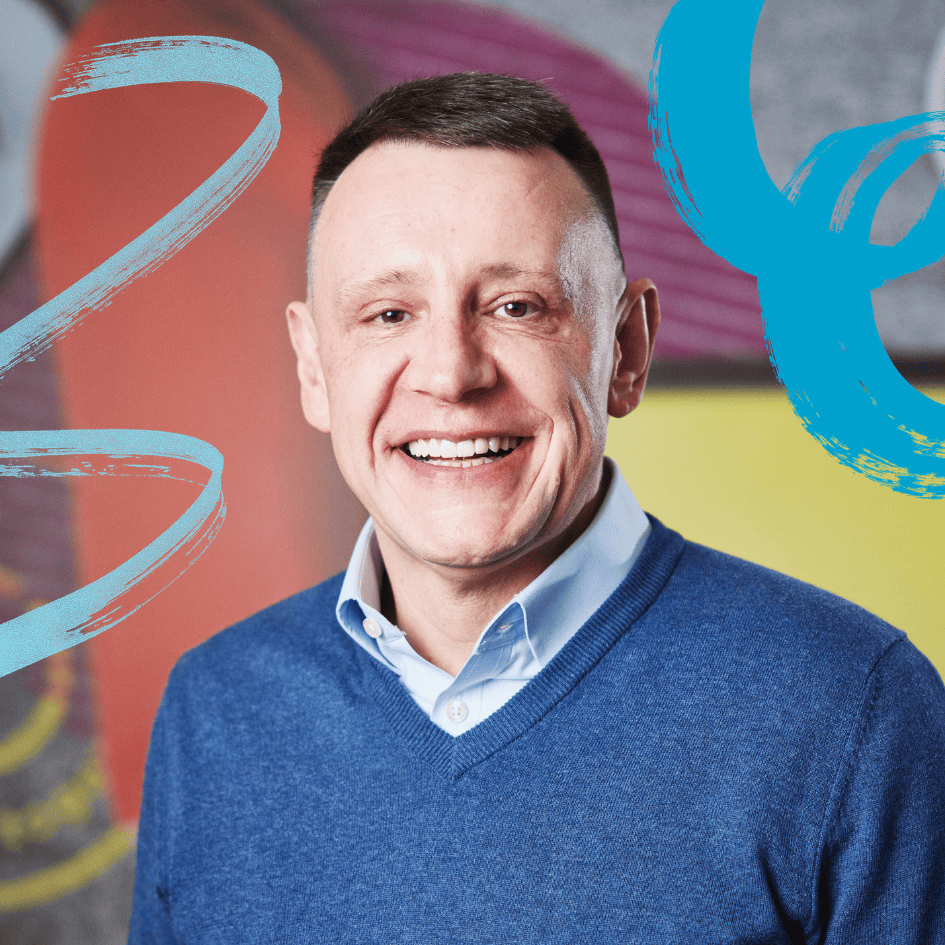Portrait of Nick Crofts, CEO of Co-op Foundation, wearing a blue sweater over a collared shirt, facing the camera against a colourful abstract background.