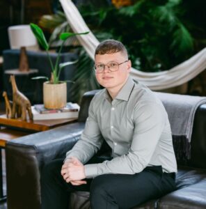 Image of Jaiden Corfield, Co-op Foundation trustee. Image shows young white male wearing a grey shirt and black trousers sitting on a sofa with plants in the background