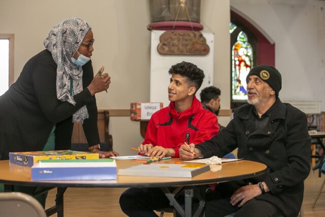 A woman speaks to a young man and and older man at a table. The young man is filling in paperwork and smiling