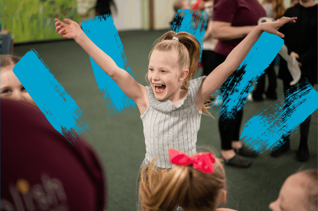 Young girl wearing a grey outfit with arms in the air, smiling excitedly.