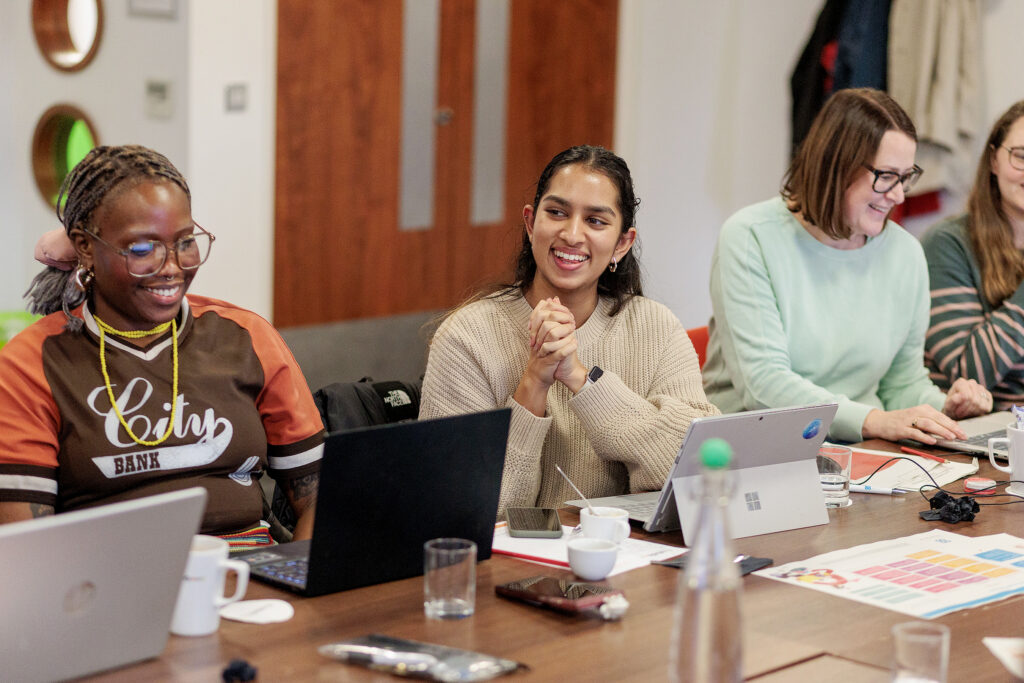 Several people sit around a table in a meeting room smiling and laughing, working on laptops with papers, cups, and bottles spread across the table.