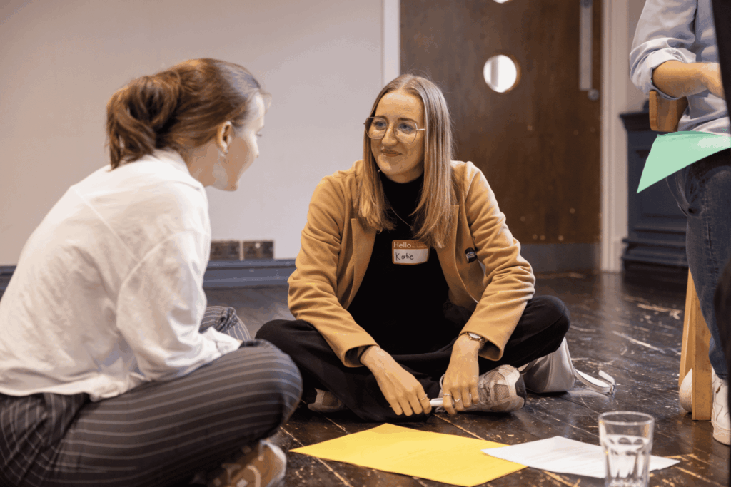 Two people sat together on the floor with pens and paper in front of them, smiling at each other.