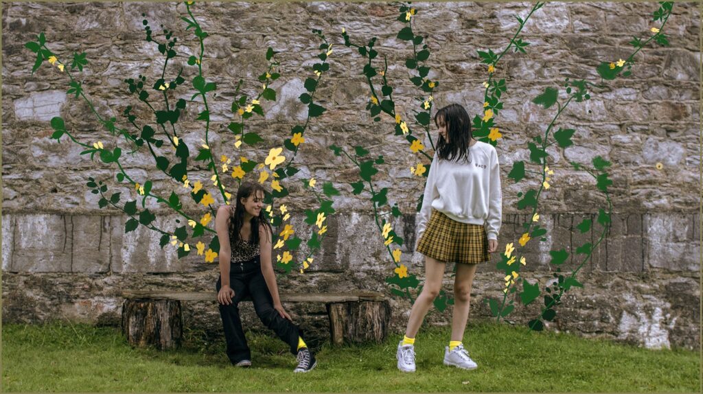 Two teenage girls pass each other in front of an old wall. They are so delighted to see they are both wearing yellow socks, that the wall spontaneously blossoms with beautiful yellow flowers.