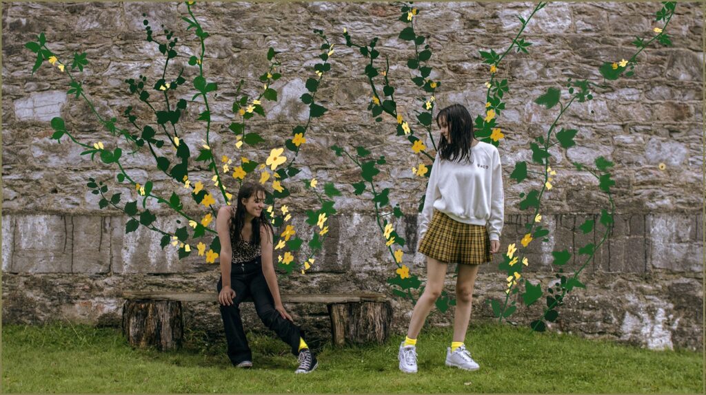 Two teenage girls pass each other in front of an old wall. They are so delighted to see they are both wearing yellow socks, that the wall spontaneously blossoms with beautiful yellow flowers.  