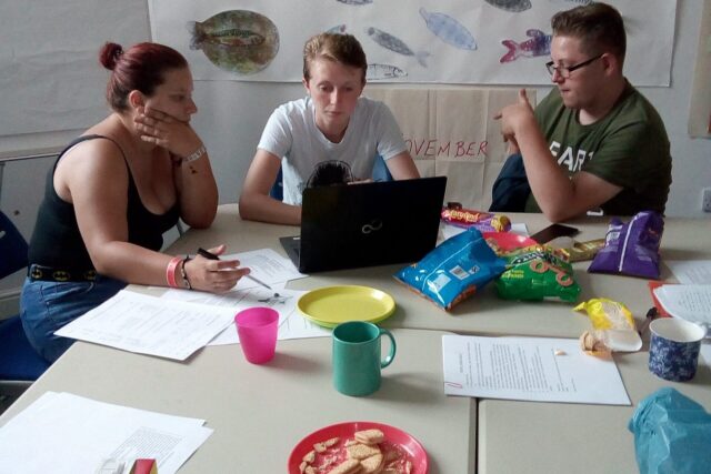 Three people sat at a table, with a laptop, papers and snacks